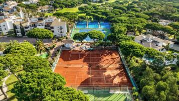 An aerial view of a residential area with a tennis court surrounded by lush greenery and white buildings with red-tiled roofs.