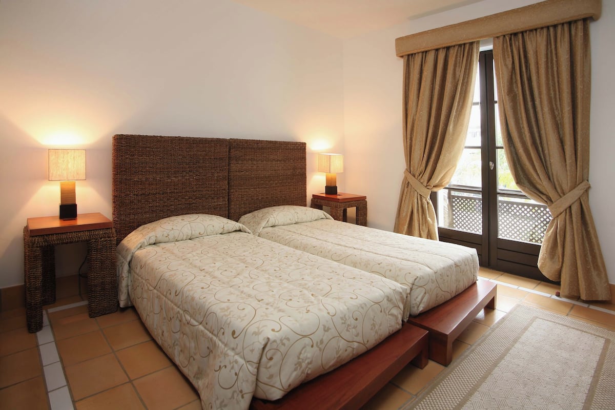 A double bed with a wooden headboard, two bedside tables with lamps, a bench at the foot of the bed, and a view through a window with curtains.