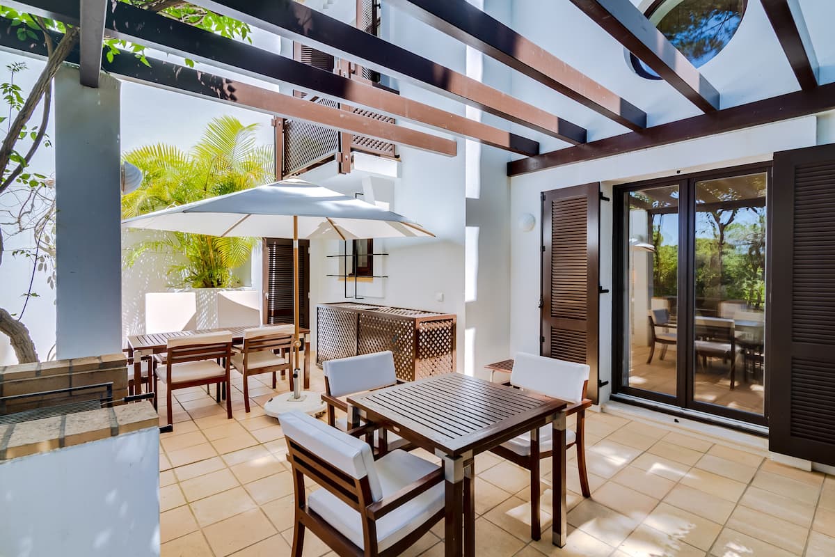 A patio area with a wooden table and chairs, a white umbrella, and a view of the outdoors.