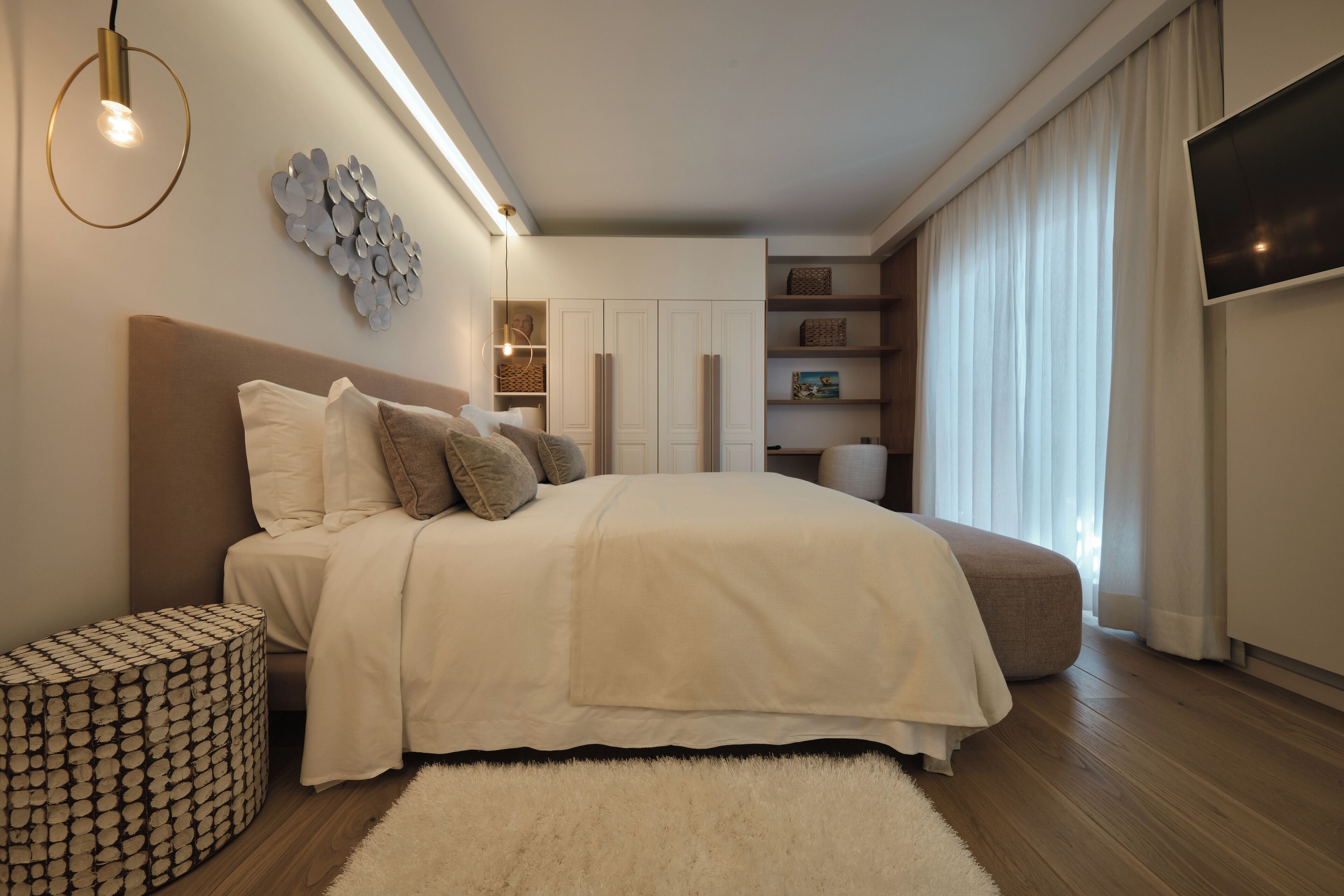Une chambre moderne avec un grand lit, un pouf rond, un téléviseur fixé au mur et des étagères intégrées.
