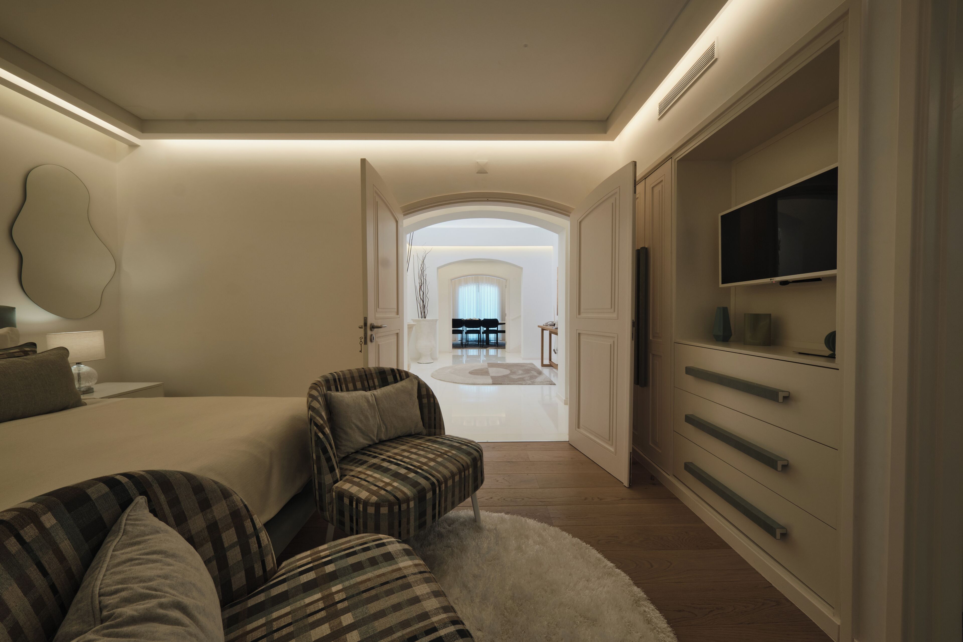 A modern bedroom with a bed, a seating area with a chair and a sofa, a TV mounted on the wall, and a view into the dining area.