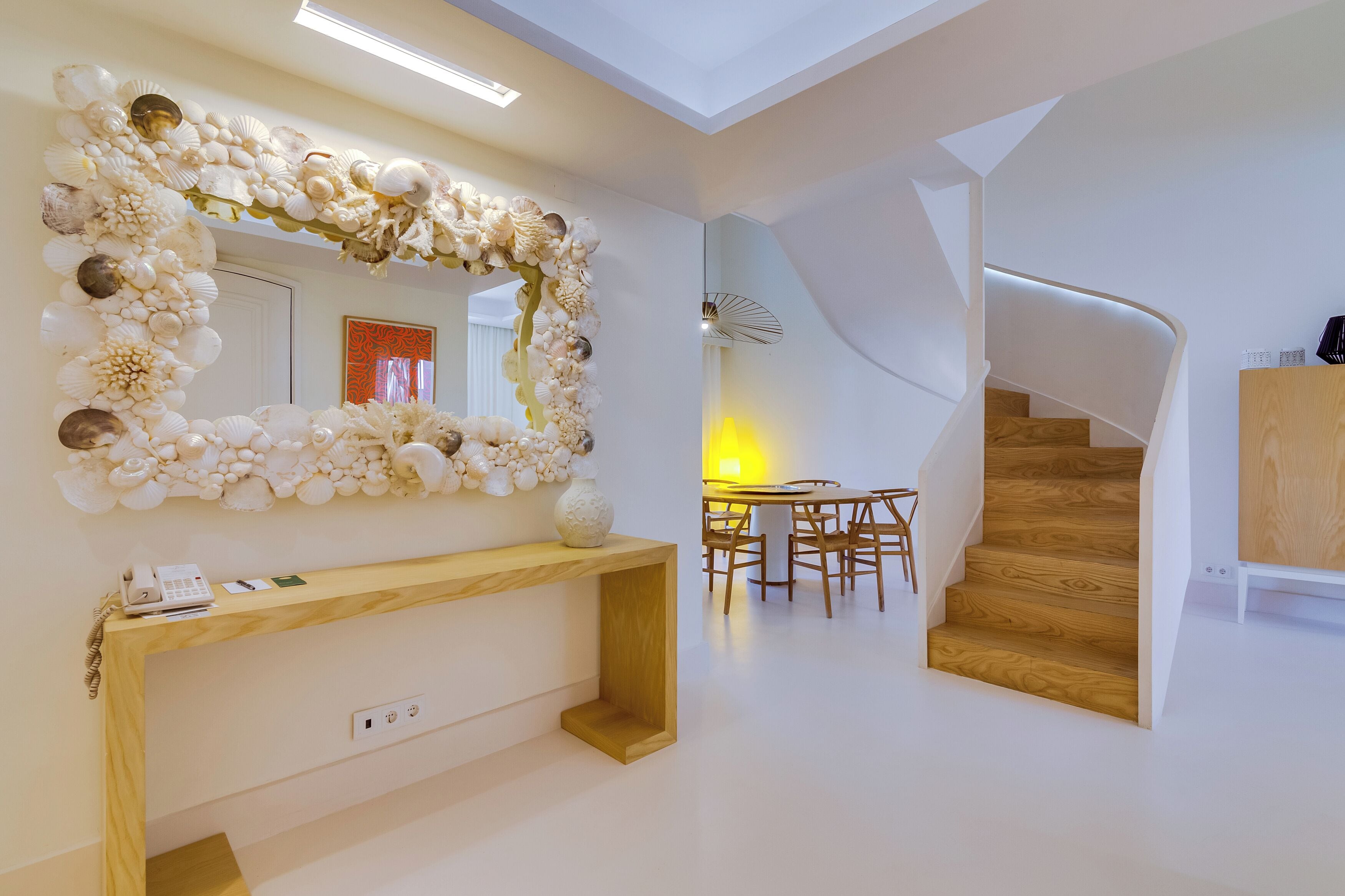 A modern interior with a wooden console table, a mirror with a shell and flower decoration, a staircase, and a dining area.