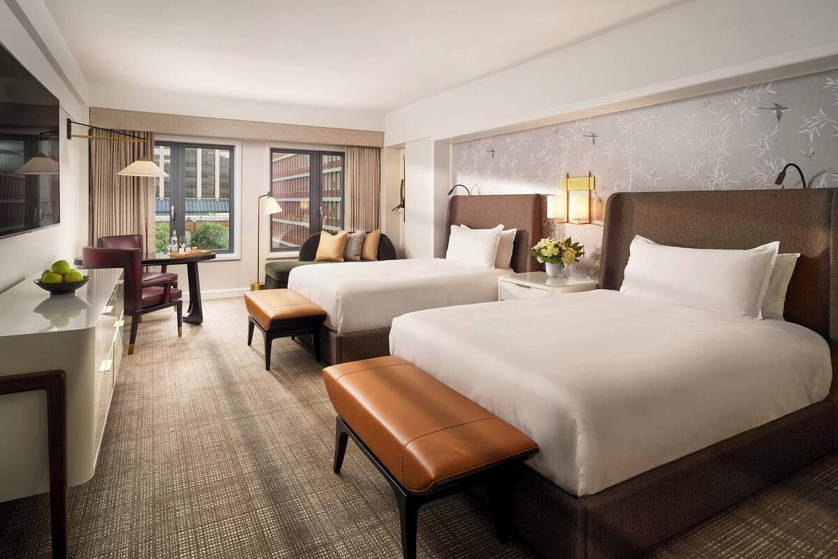 A hotel room with two beds, a desk, a chair, a bench, and a view of the outside.