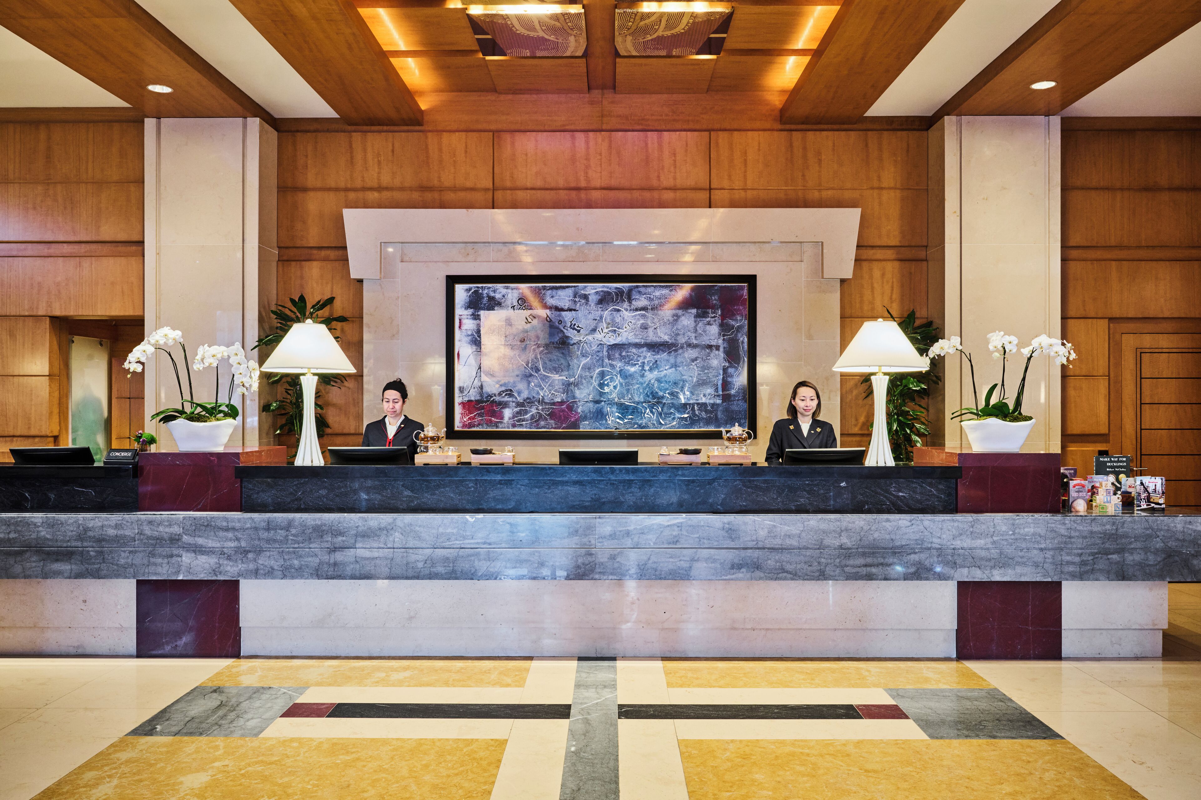 A hotel reception desk with two receptionists, a large abstract painting above, and potted orchids on either side.