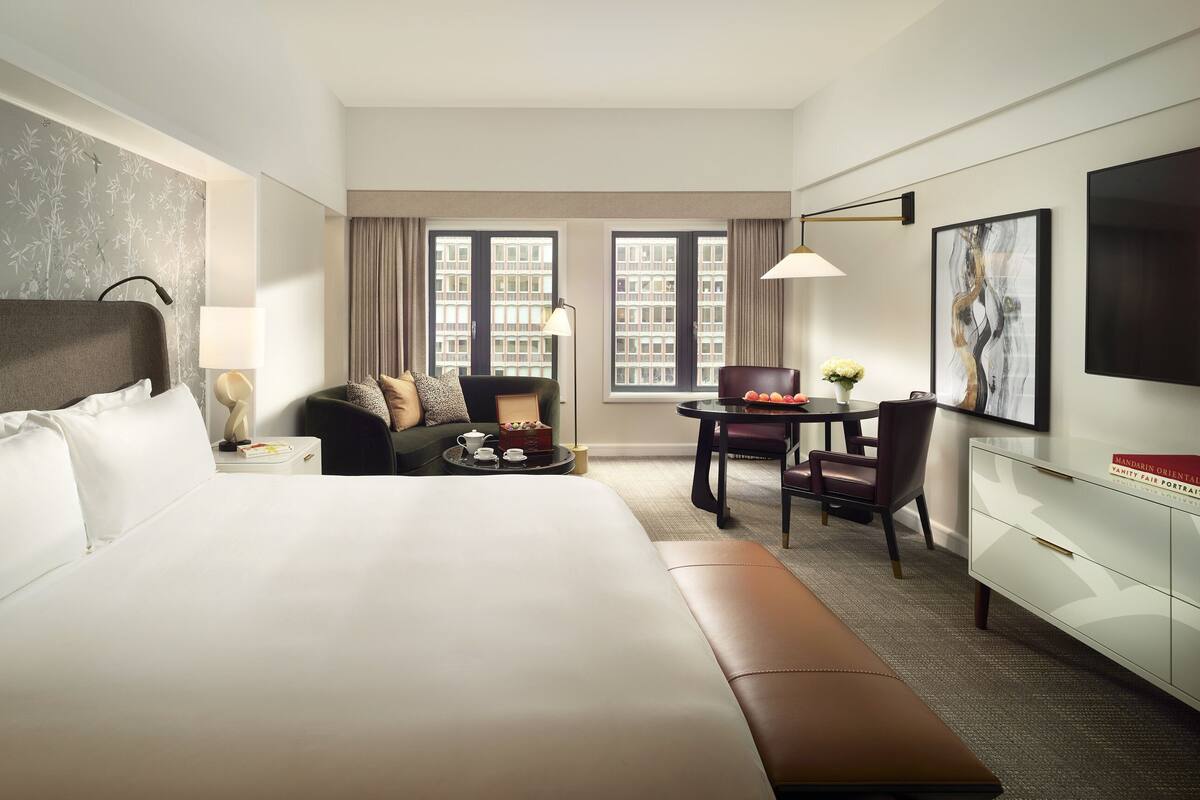 A hotel room with a large bed, a sitting area with a table and chairs, a desk, a lamp, and a large window with a city view.