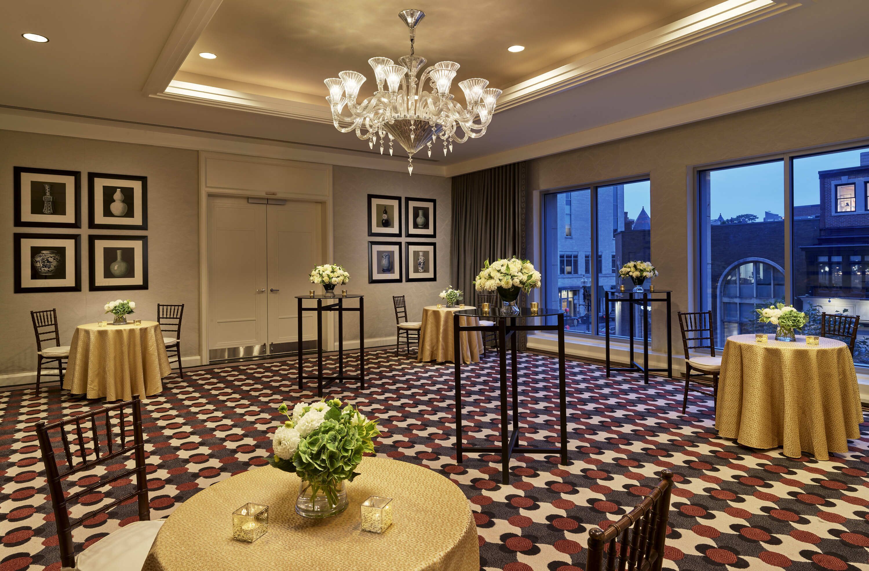 A hotel room with a patterned carpet, multiple tables with round tables and square tables, a chandelier, and framed pictures on the walls.