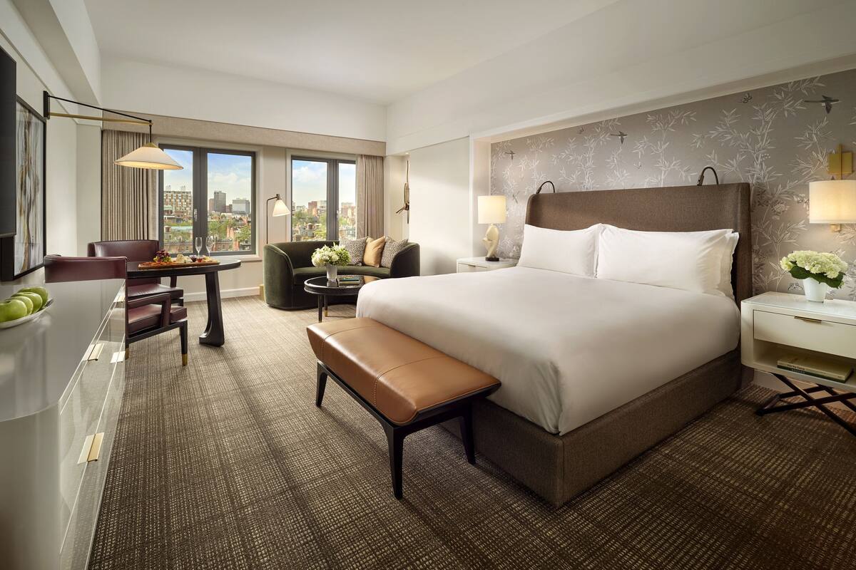 A hotel room with a large bed, a desk with a chair, a bench, and a view of the city through large windows.