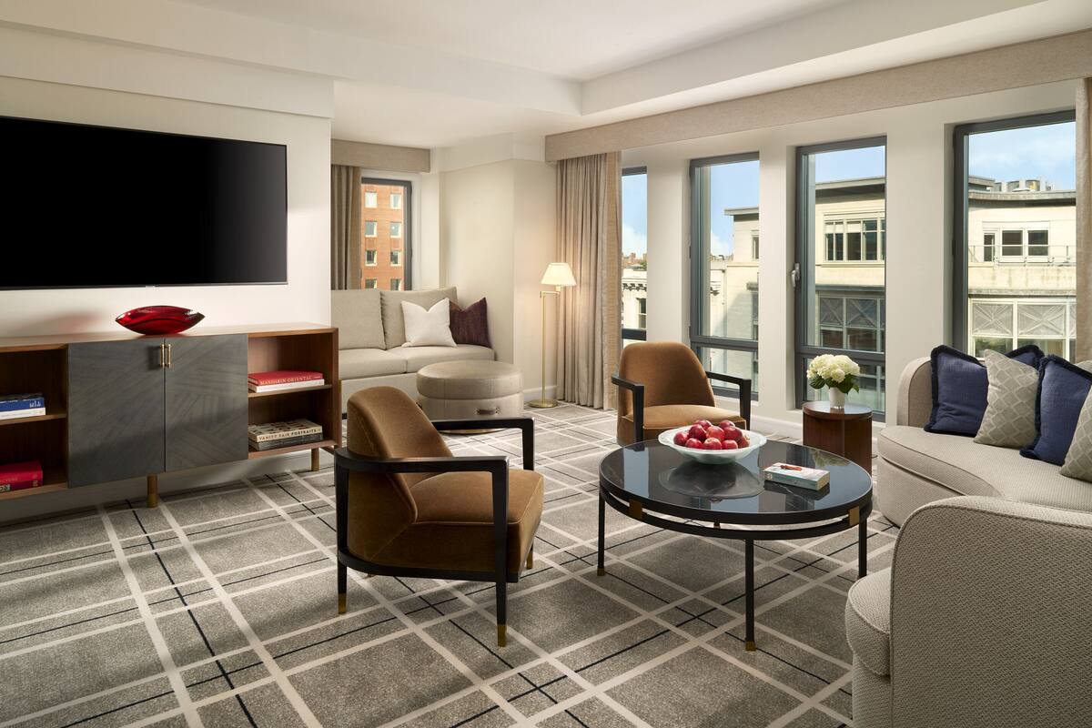 A modern hotel room with a flat-screen TV, a sofa, armchairs, a coffee table, and a view of the city through large windows.