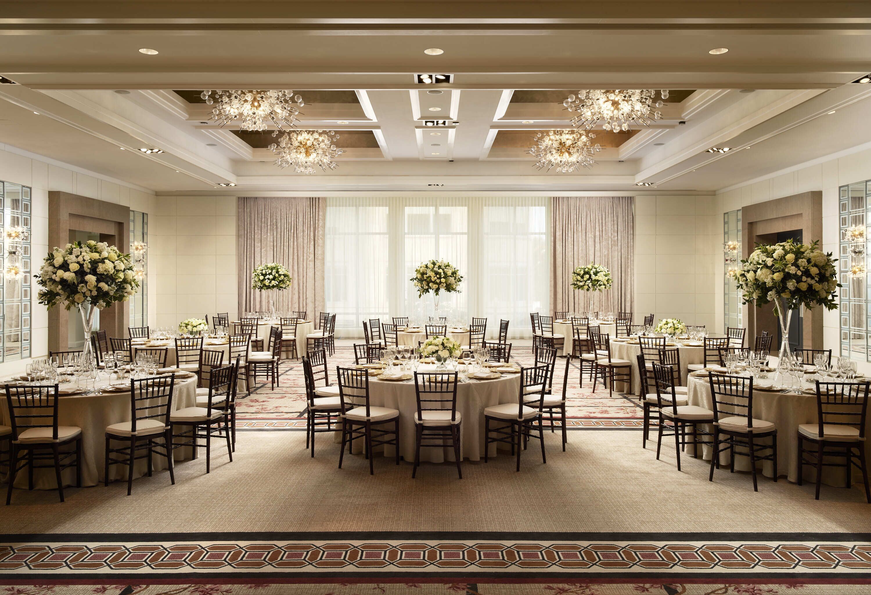 A spacious banquet hall with multiple round tables, each set with chairs, surrounded by large windows and adorned with floral arrangements.