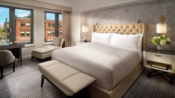 A hotel room with a large bed, a desk, a chair, a bench, and a view of buildings through the window.