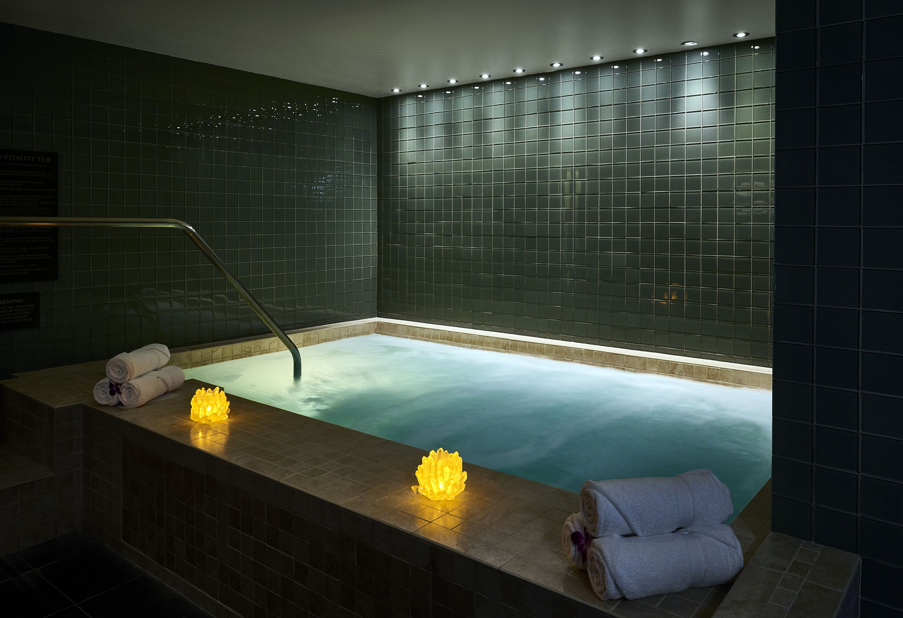 Zona de spa con bañera de hidromasaje, paredes con azulejos y una pieza decorativa iluminada en el borde.