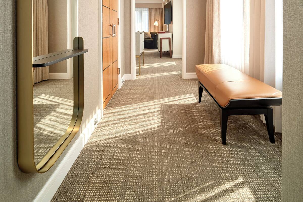 A hotel corridor with a bench, a mirror, and a view into a room with a bed and a TV.