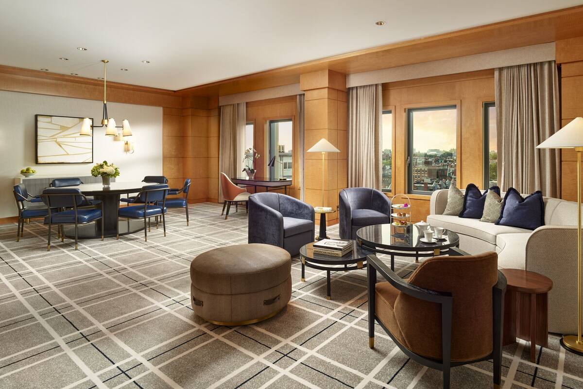 A spacious hotel room with a large window offering a city view, a cozy seating area with a sofa and armchairs, a dining table with chairs, and a small side table with a lamp.