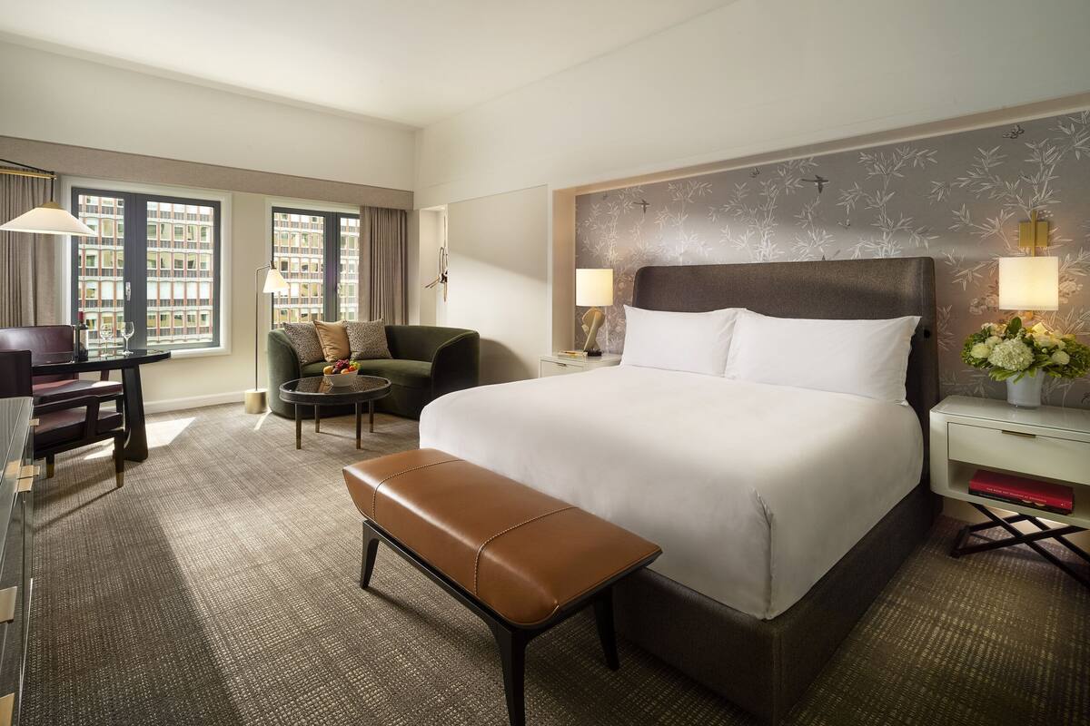 A hotel room with a large bed, a desk, a chair, a bench, and a view of buildings through the window.