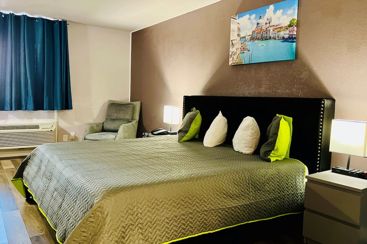 A hotel room with a bed, a chair, a nightstand, a lamp, and a painting of a cityscape.