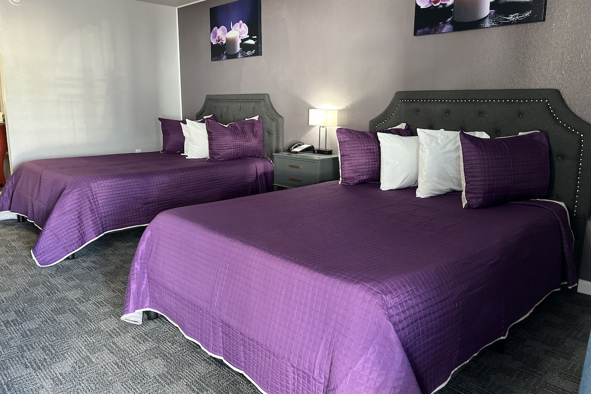 A hotel room with two beds, each with purple bedding and white pillows, a nightstand with a lamp, and a wall-mounted picture of orchids.