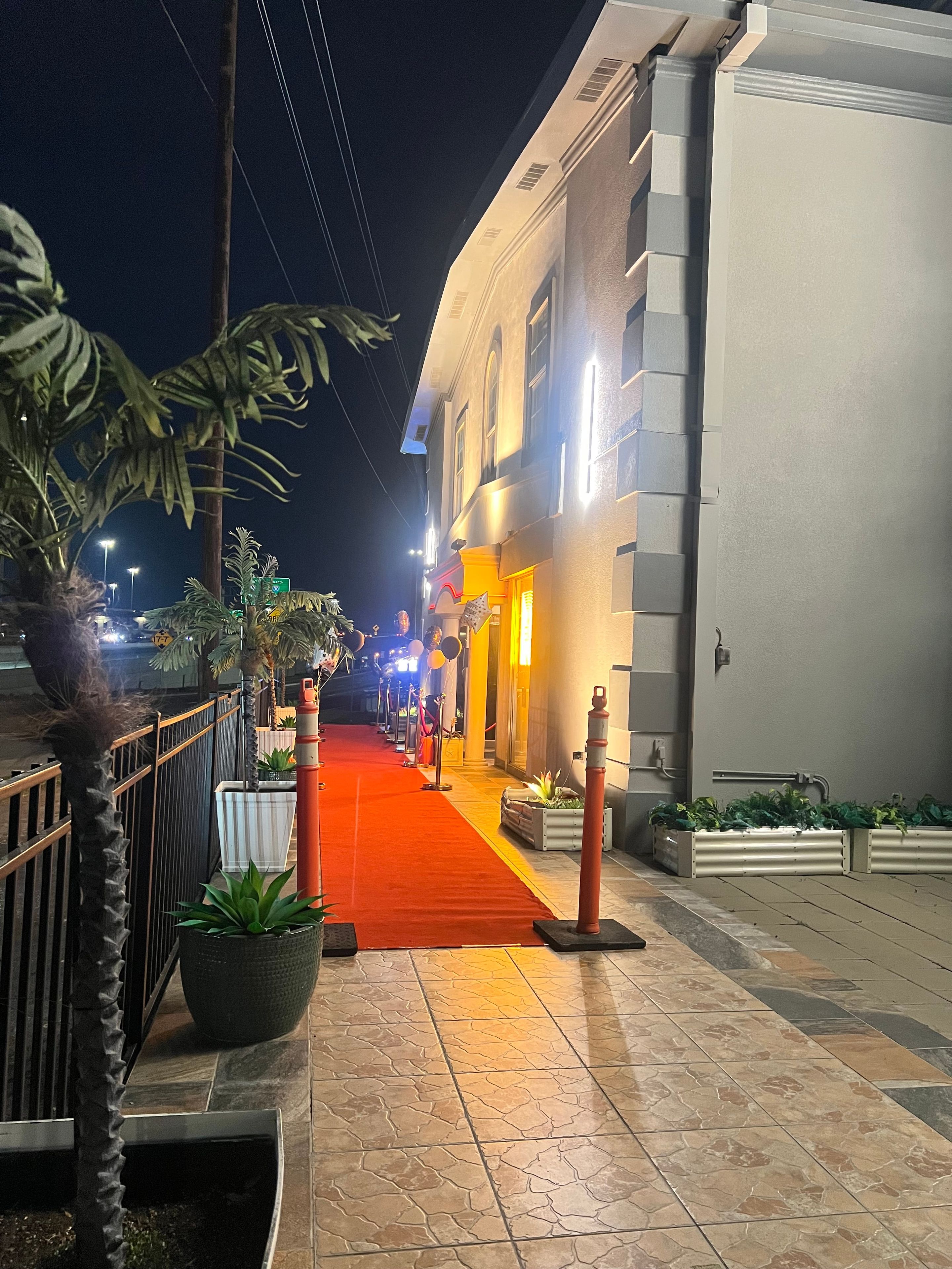 Vista nocturna de una calle con una alfombra roja que conduce a un edificio de fachada blanca y varias ventanas.