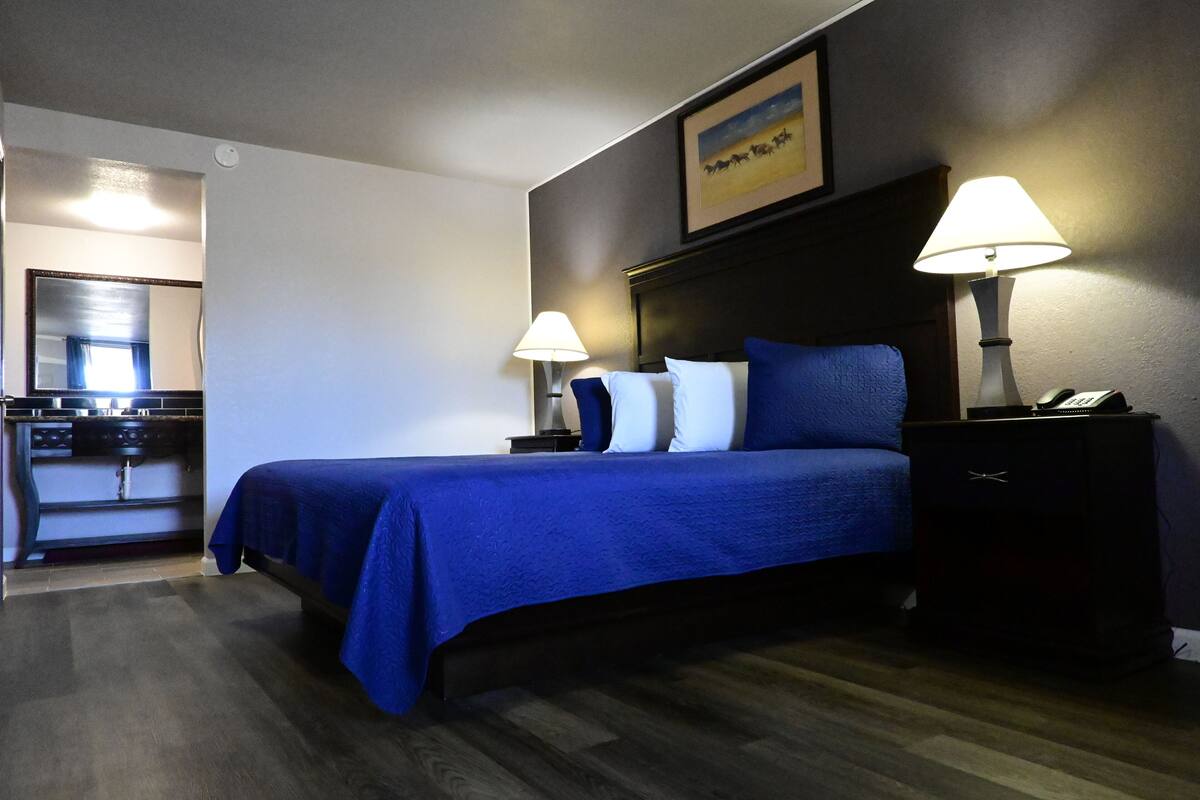A hotel room with a bed, two bedside lamps, a nightstand, a framed picture on the wall, and a small table with a lamp.