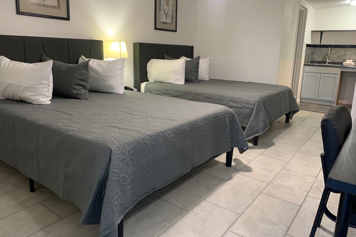A hotel room with two beds, a desk, a chair, a lamp, and a kitchenette.