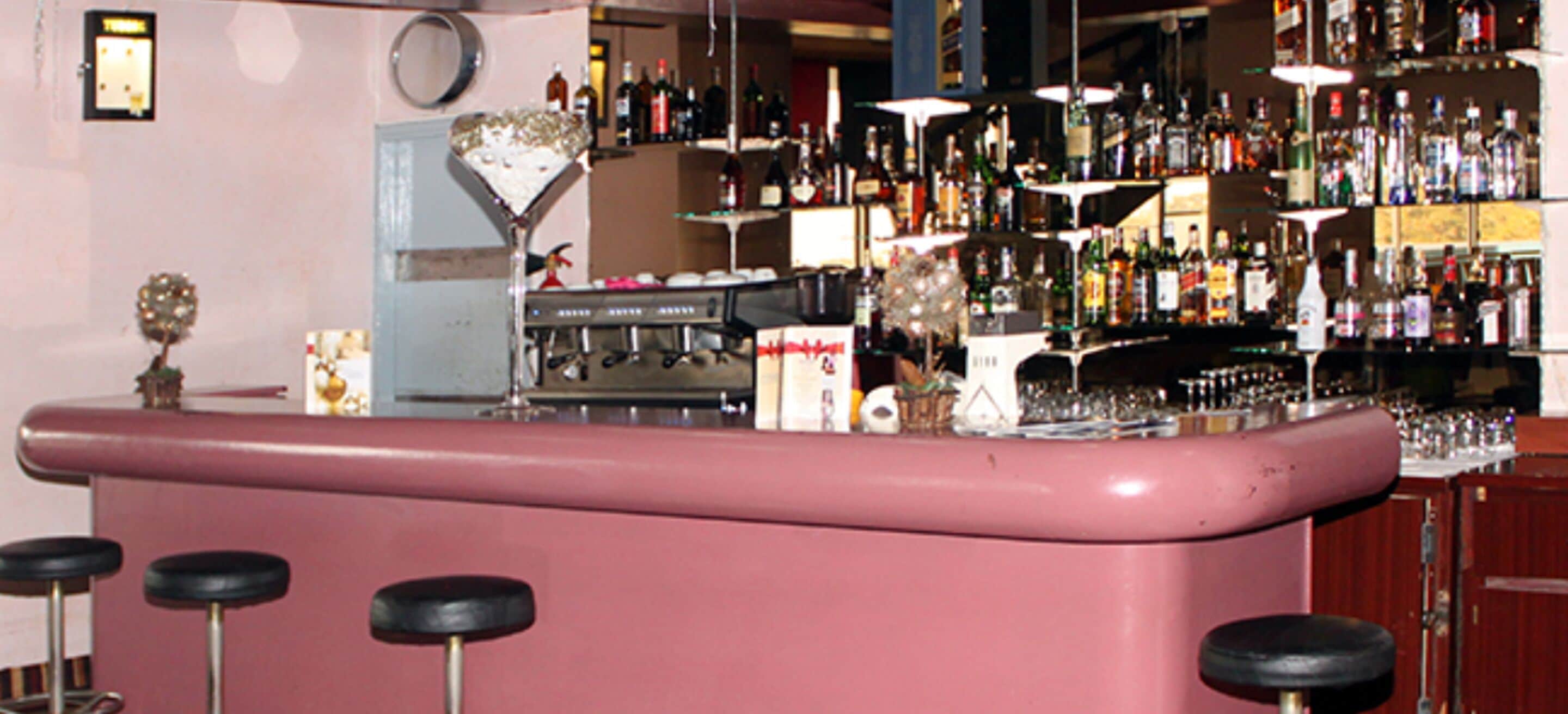 Een bar met een roze toonbank, krukken en planken vol verschillende flessen.