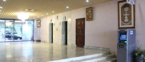 Un vestíbulo con suelo de mármol, un mostrador de recepción, un cajero automático y una puerta con panel decorativo.