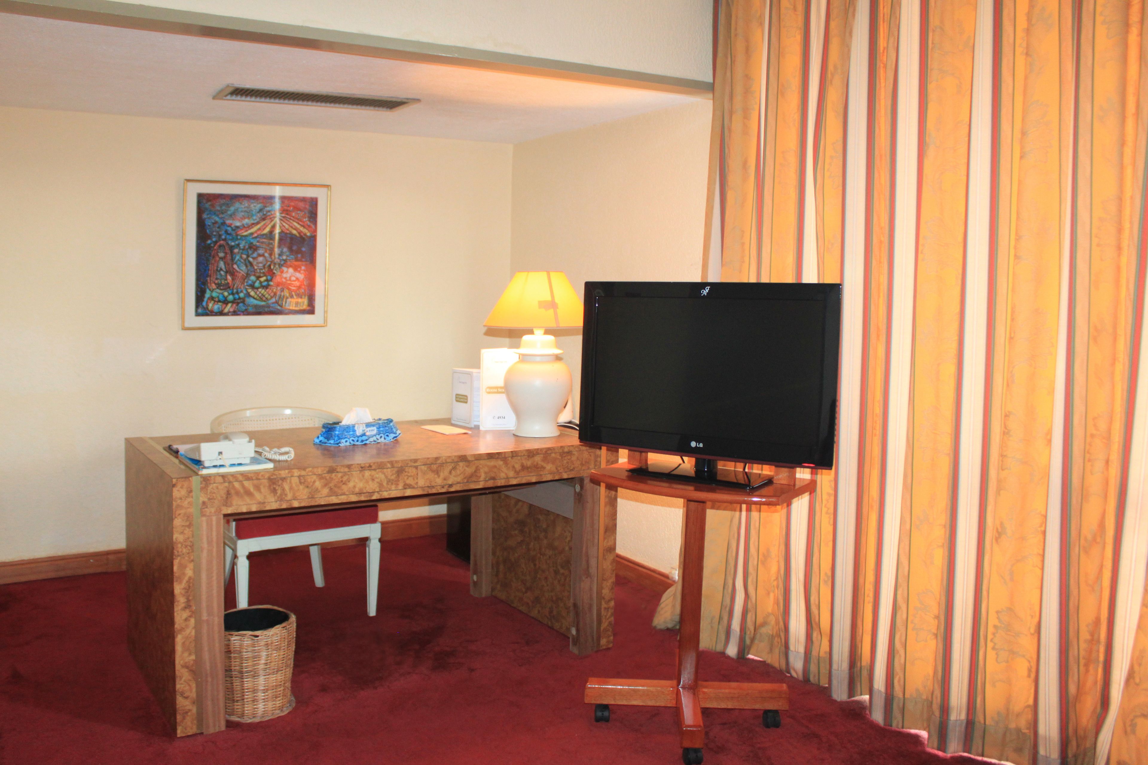 Ein Hotelzimmer mit einem Holztisch, einem Fernseher auf einem Standfuß, einer Lampe, einem Mülleimer und einem gerahmten Bild an der Wand.