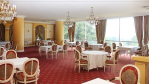 Ein formeller Speisesaal mit Tischen, Stühlen, einer Kronleuchter und großen Fenstern mit Vorhängen.
