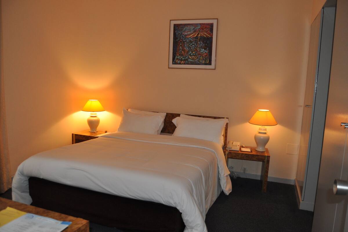 A hotel room with a bed, two bedside lamps, a small table with a lamp, a painting on the wall, and a door.