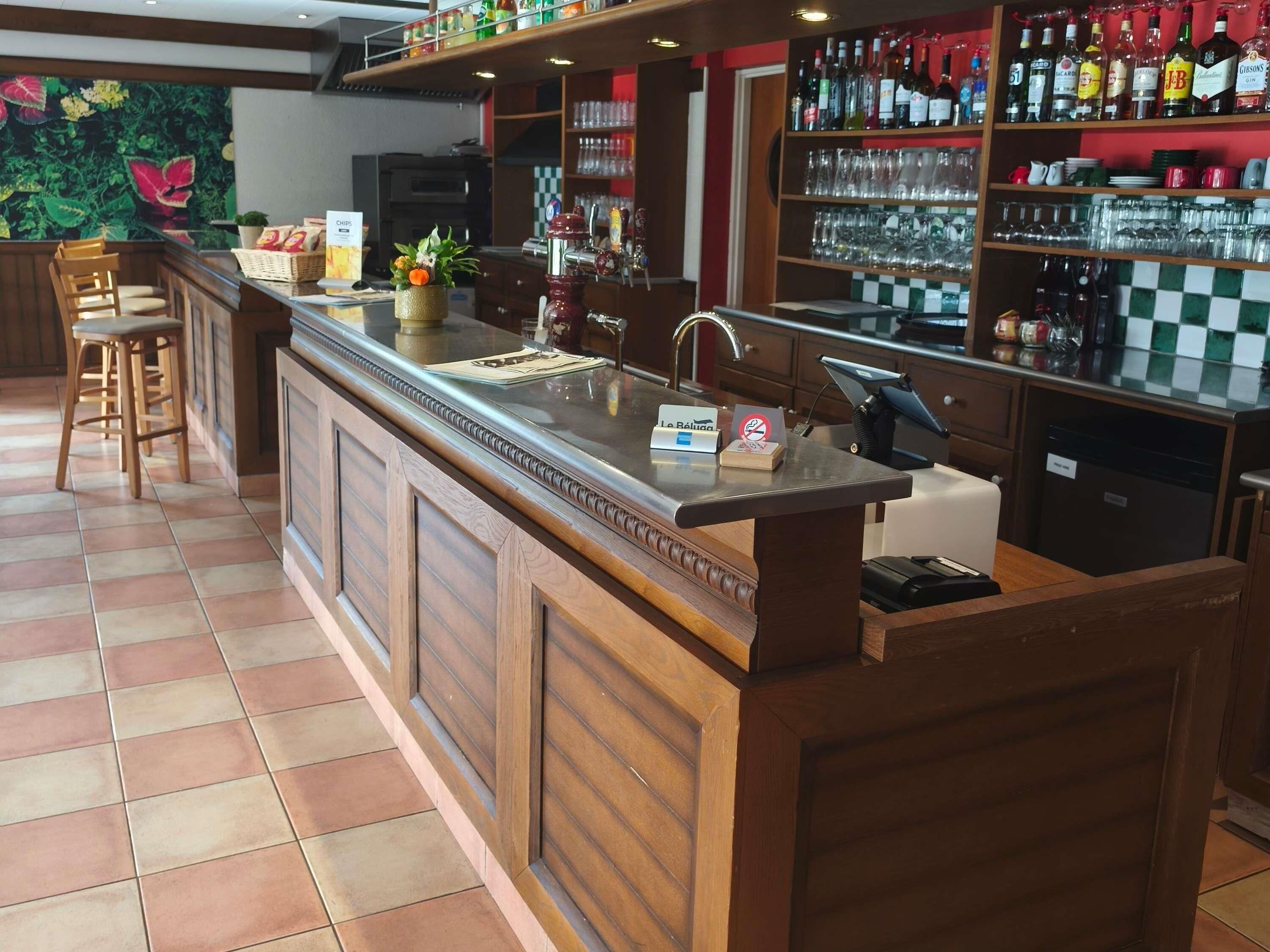 A bar area with a counter, stools, a cash register, and a shelf with bottles and glasses.