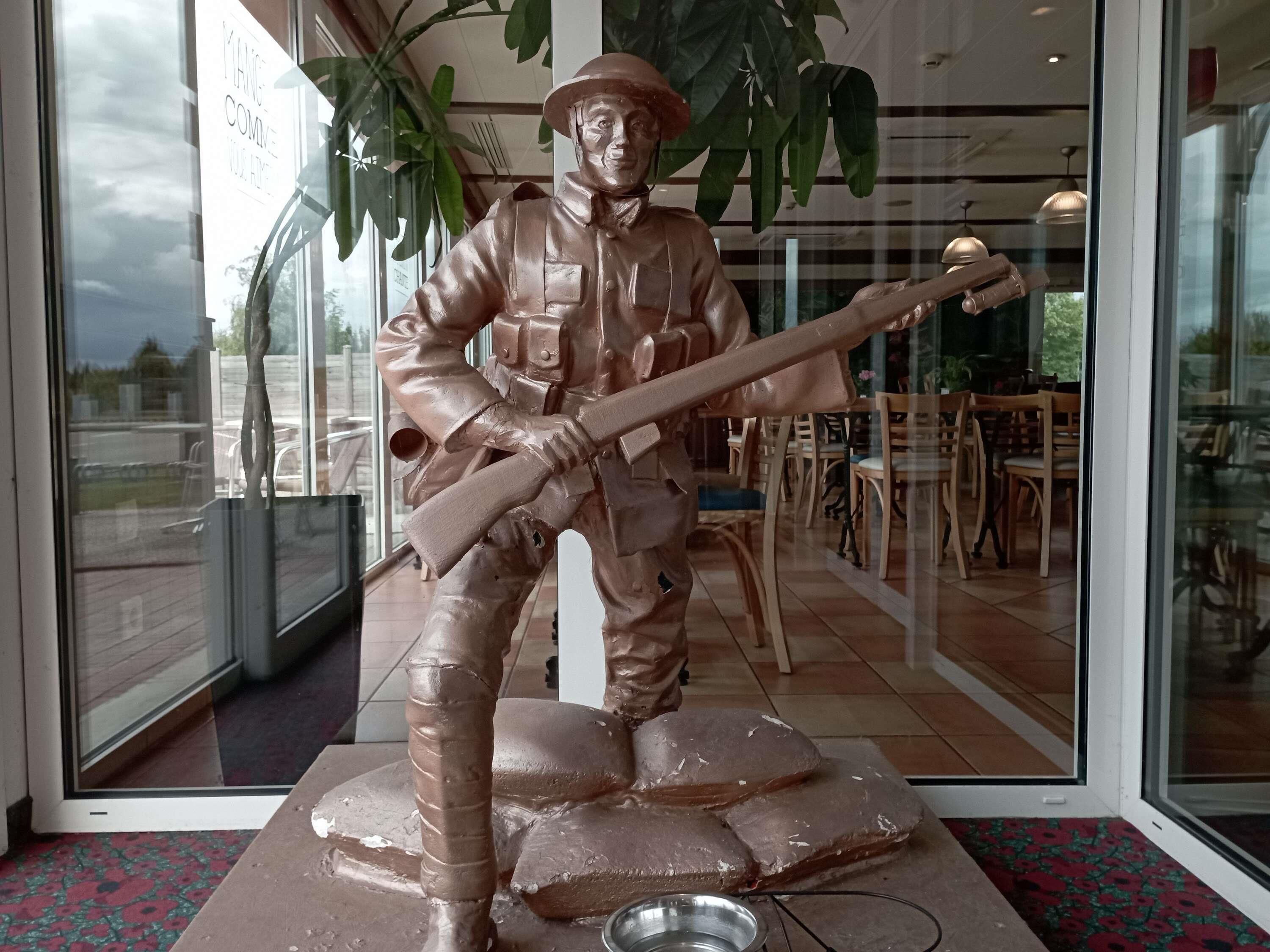 A bronze statue of a soldier holding a rifle, placed on a pedestal in a room with large windows, wooden furniture, and a tiled floor.