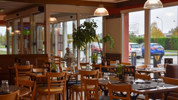 Interior de restaurante con mesas y sillas de madera, plantas en macetas y grandes ventanales con vista a coches aparcados.