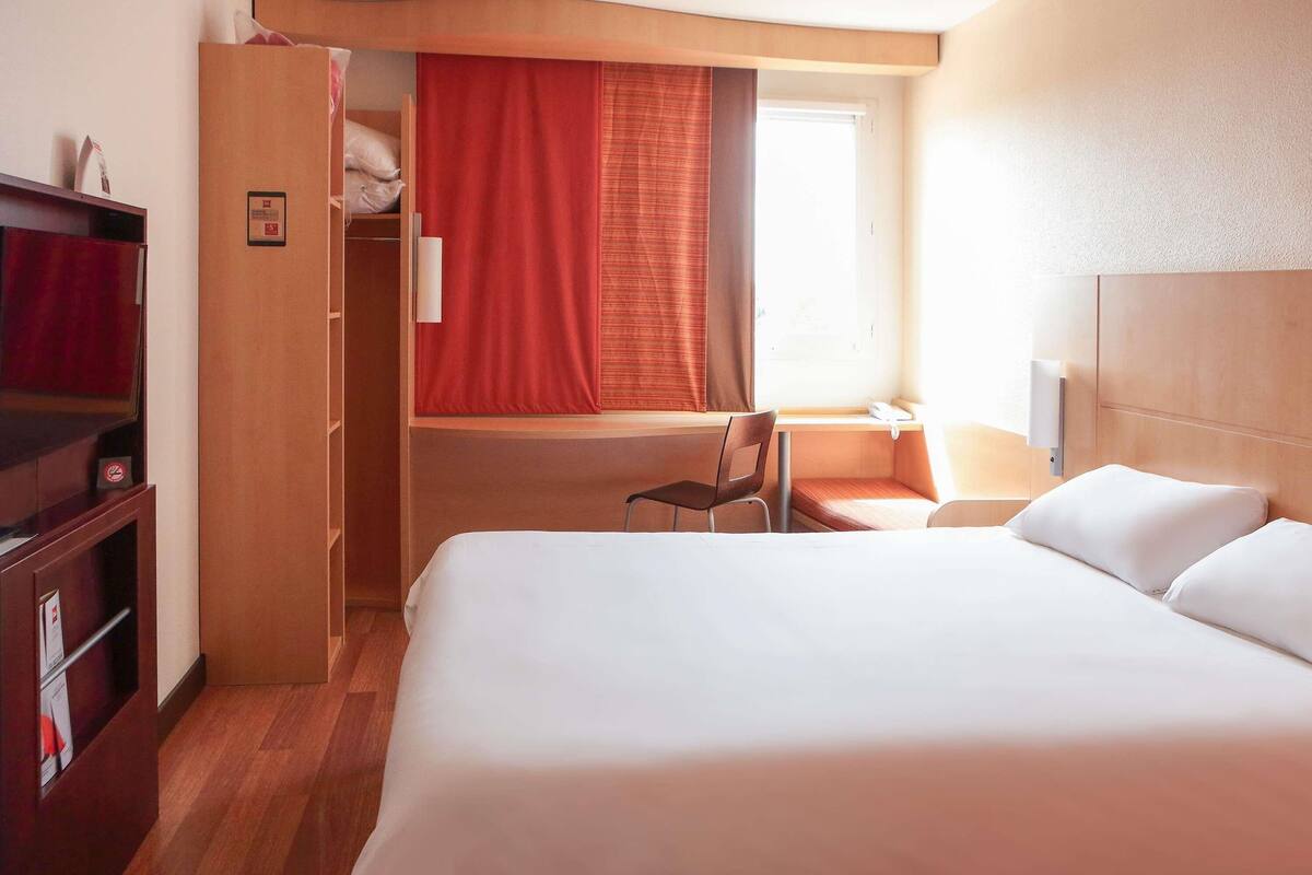Ein Hotelzimmer mit Bett, Schreibtisch mit Stuhl, Kleiderschrank und einem Fenster mit Vorhängen.