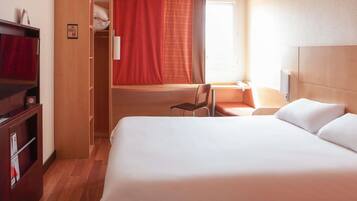 A hotel room with a bed, a desk with a chair, a wardrobe, and a window with curtains.