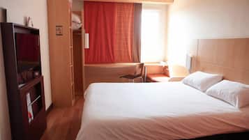 Une chambre d’hôtel comprenant un lit, un bureau, une chaise, une armoire et une fenêtre avec des rideaux.