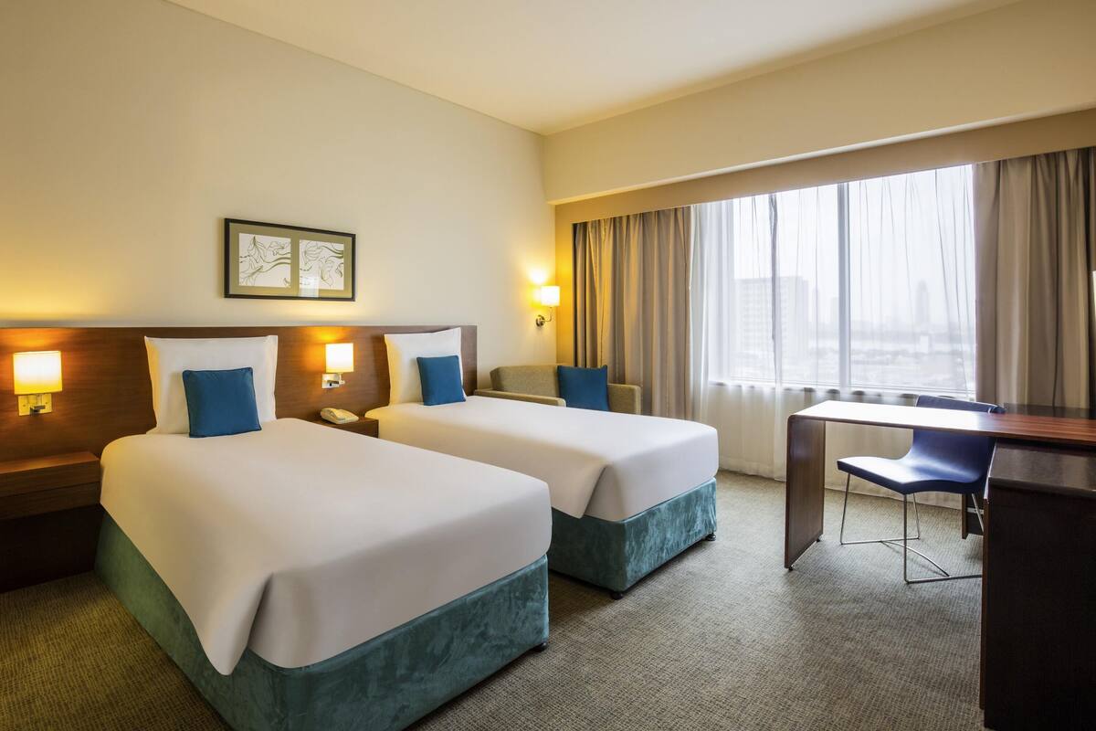 Une chambre d’hôtel avec deux lits, un bureau et une chaise. On y trouve deux lampes de chevet et une fenêtre avec des rideaux.