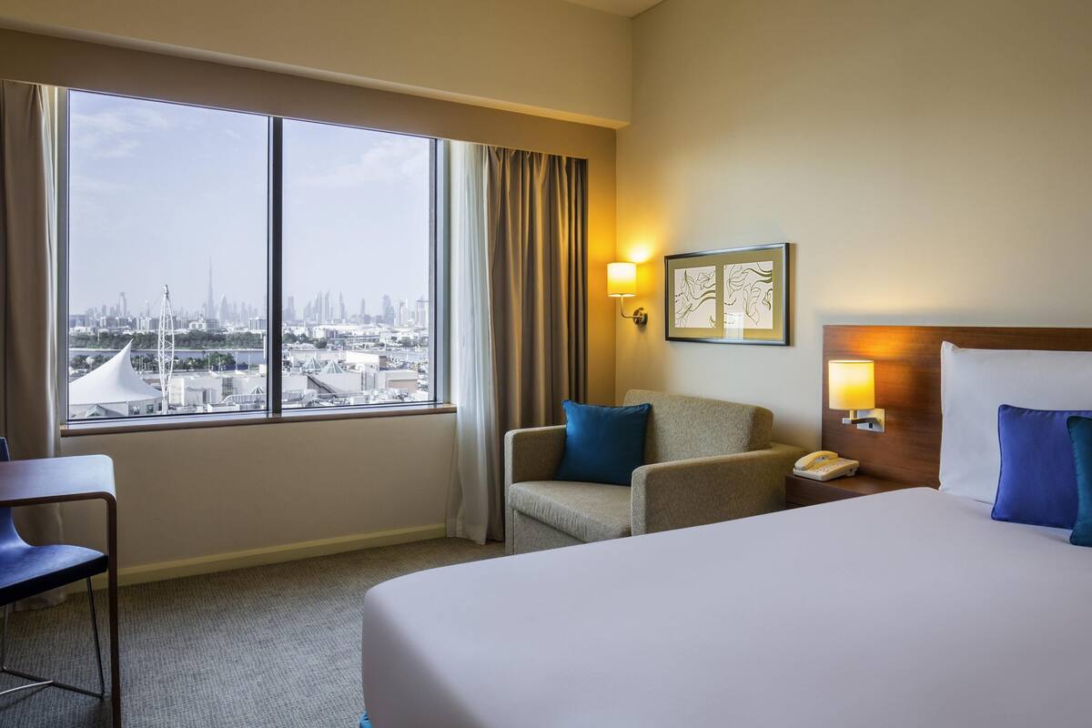 Une chambre d’hôtel équipée d’un lit, d’une chaise, d’un bureau, d’un téléphone et offrant une vue sur le paysage urbain grâce à de grandes fenêtres.
