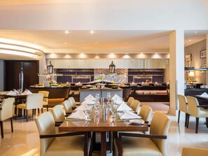 A hotel dining area with a long table set for a meal, a kitchen in the background, and a modern interior design.
