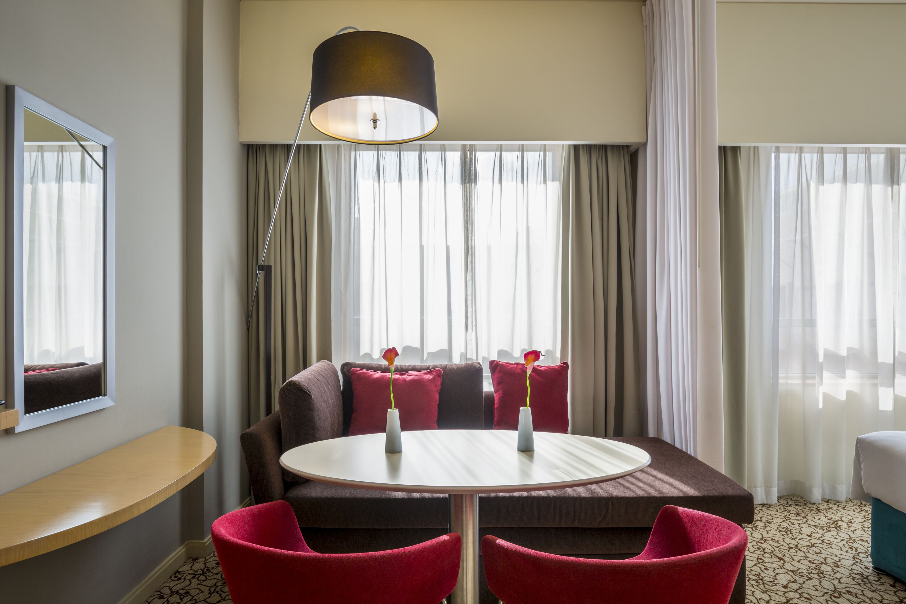 A hotel room with a round table, two red chairs, a sofa with red cushions, a large window with sheer curtains, and a floor lamp.