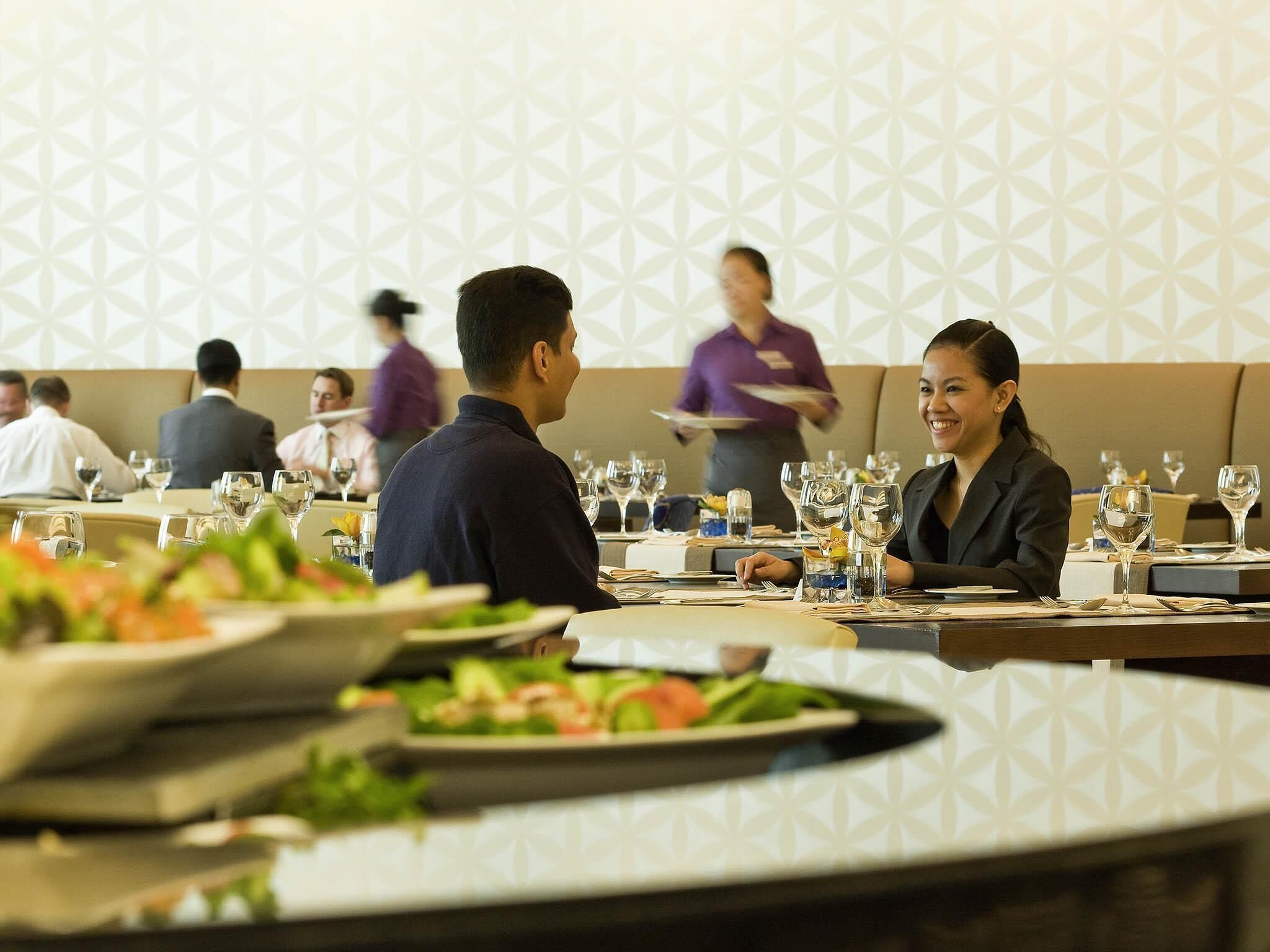Une salle de restaurant avec des tables dressées, un serveur en arrière-plan et un mur orné d’un motif.