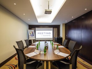 Kokoushuone, jossa on suuri pöytä, tuoleja, projektori, valkokangas, jossa näkyy teksti ”KOKOUS ATNOVOTEL”, ja fläppitaulu.