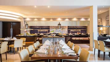 A hotel dining area with a long table set for a meal, a kitchen in the background, and a modern interior design.