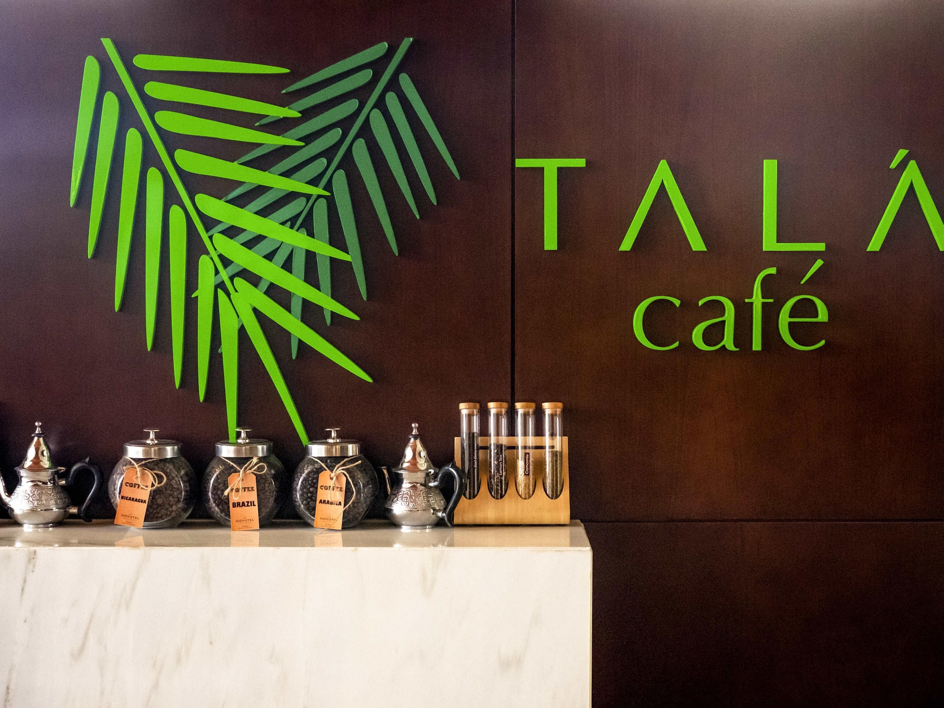 Un mur orné d’une décoration représentant des feuilles vertes, sur lequel on peut lire en lettres vertes l’inscription « TALÁ café ». Plusieurs distributeurs de café, chacun portant une étiquette, sont disposés sur le comptoir en contrebas.