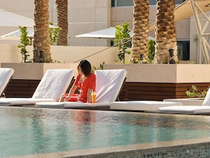 A poolside area with lounge chairs, palm trees, and a person in a red dress. - Novotel Deira Creekside Dubai (Dubai)