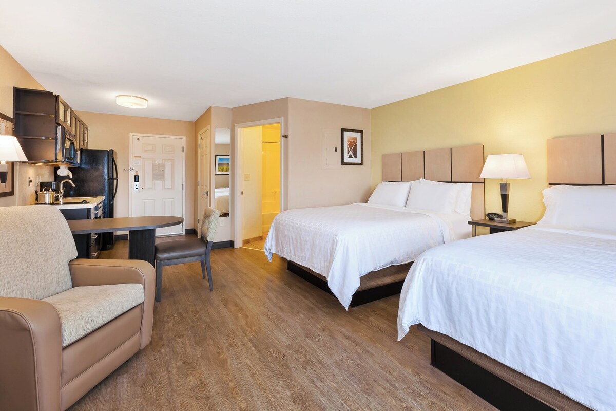 A hotel room with two beds, a sofa, a desk with a chair, a kitchenette, and a door leading to another room.