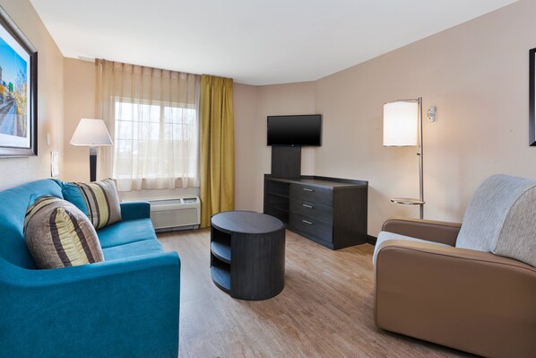 A modern living room with a blue sofa, a brown sofa, a flat-screen TV, a lamp, and a large window with curtains. - Candlewood Suites Rocky Mount by IHG (Rocky Mount)