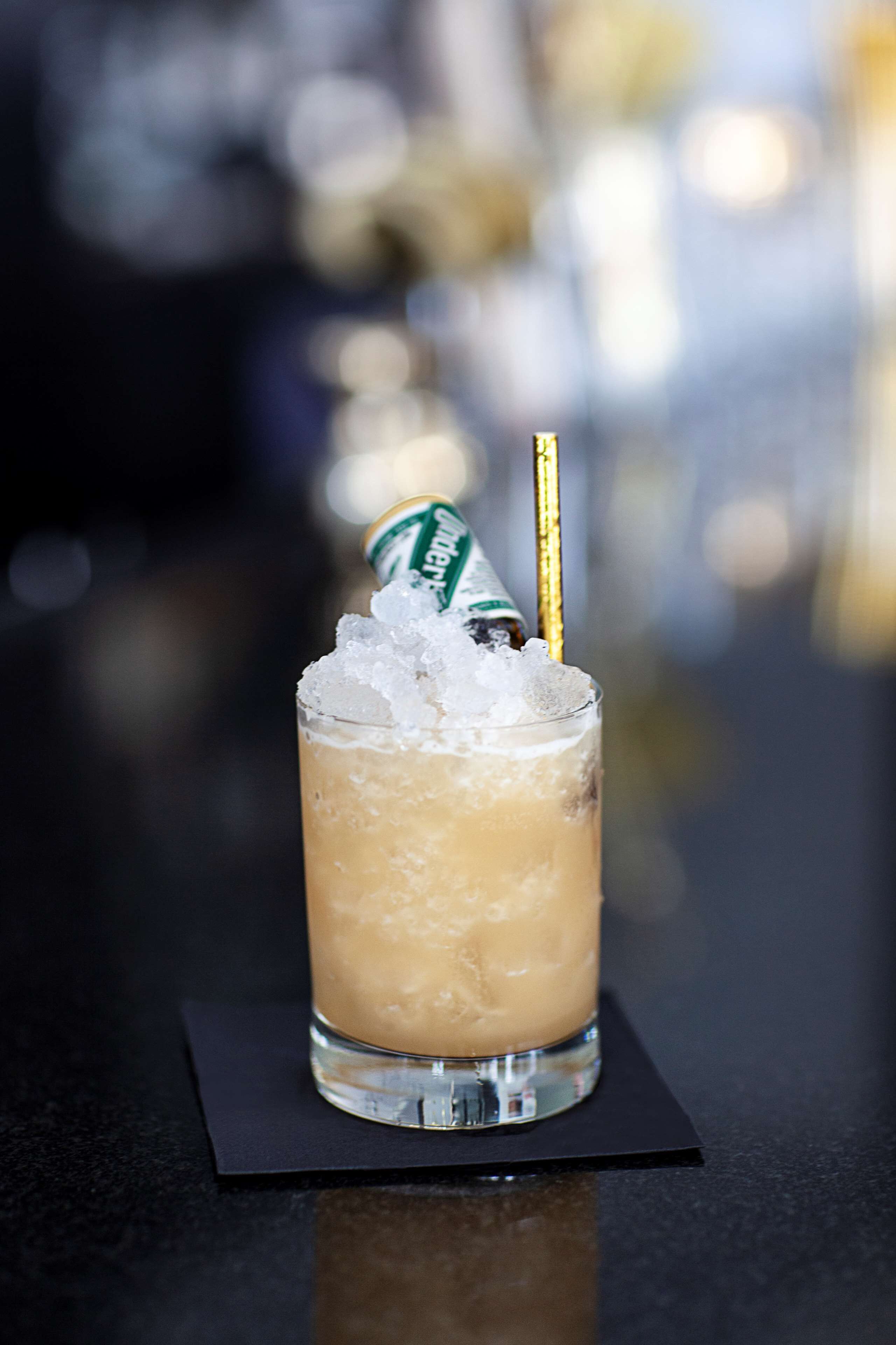 A tall glass with a straw, filled with a light-colored beverage and ice cubes, placed on a black coaster.