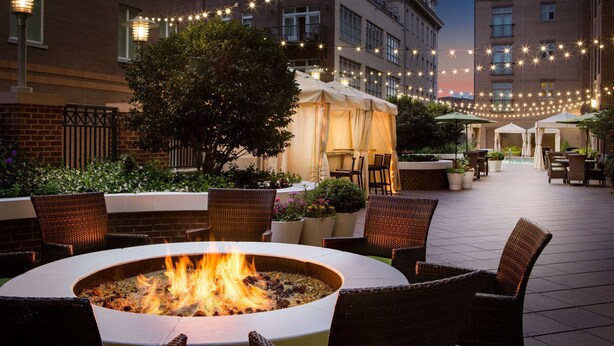 An outdoor patio with wicker furniture, a fire pit, string lights, and a building in the background. - ANDAZ SAVANNAH, BY HYATT (Savannah)