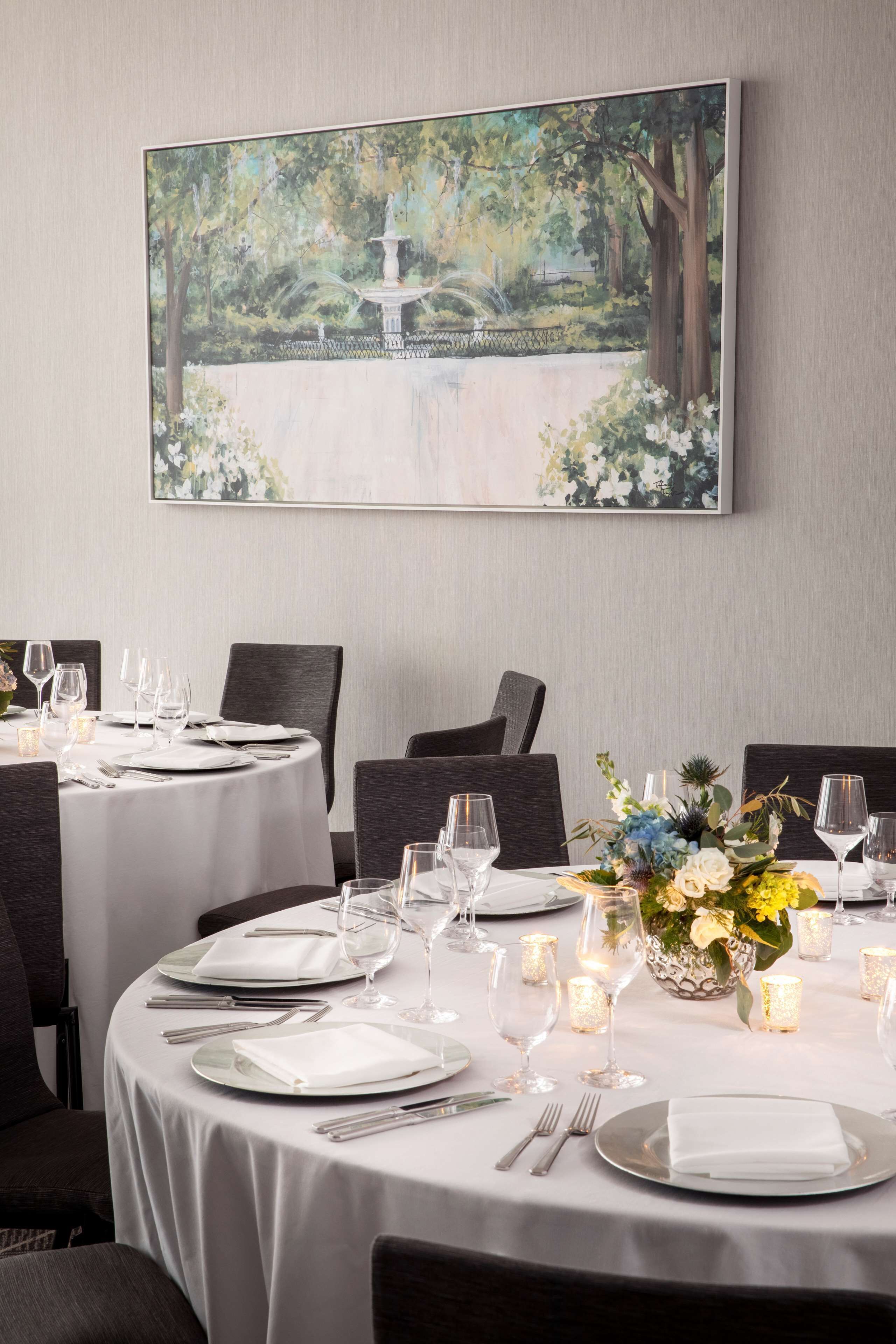 A dining area with round tables set for a meal, a large painting of a garden on the wall, and a centerpiece with flowers and candles.