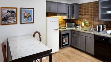 Una cocina moderna con armarios grises, un refrigerador blanco, un microondas y una cafetera. Hay una mesa de comedor con sillas y dos cuadros en la pared.