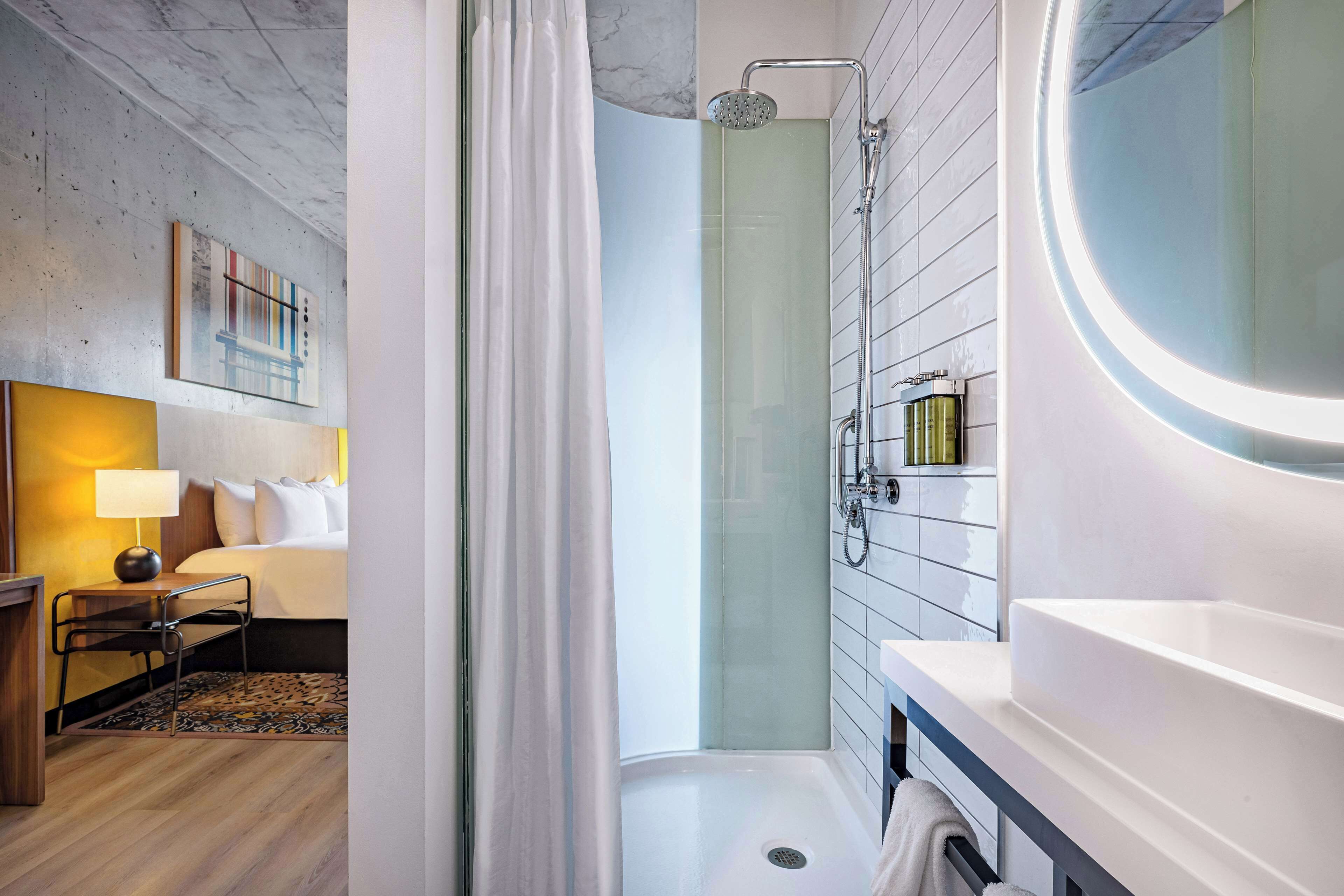 Un baño de hotel moderno con cabina de ducha de vidrio, un espejo redondo y un lavamanos blanco.